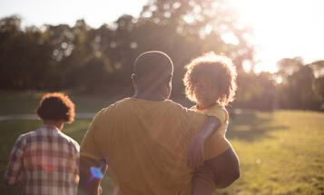 Backlit image of a Black family walking towards the setting sun across an open lawn with a toddler looking over their father's shoulder toward the camera