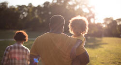 Backlit image of a Black family walking towards the setting sun across an open lawn with a toddler looking over their father's shoulder toward the camera