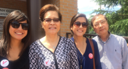 A smiling Vietnamese family stands together outdoors, with two women wearing "Vietnamese American Votes" stickers, symbolizing their participation in the voting process. The image reflects pride and unity, emphasizing the role of community support in encouraging voter participation.