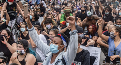 Image accompanying an opinion article titled 'Progressives Won’t Help the Working Class by Abandoning Marginalized Groups' by Taifa Butler and Carol Lautier, published by Demos. It shows a crowd of demonstrators sitting closely together, many wearing masks and raising fists in a sign of solidarity.