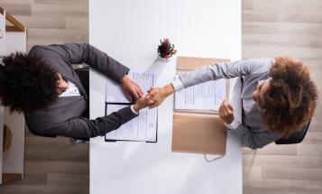Two people shaking hands across a table in an interview