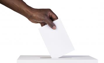 A dark-skinned hand is about to place a slip of paper in a white ballot box