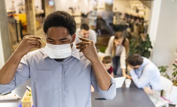 Black worker putting on mask with people waiting in the background