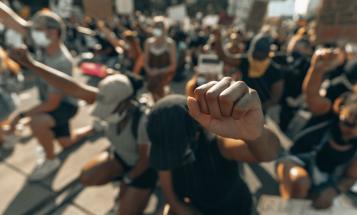 Black, brown, and white protestors knee with their heads down and their fists in the air