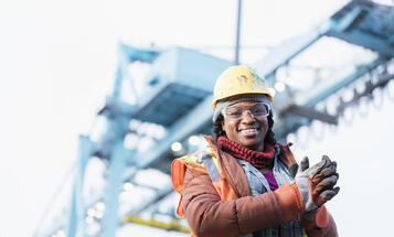 Smiling Black construction worker with a yellow hard hat and safety vest in front of a construction crane