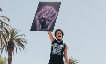 Black woman at a protest, holding up a sign of a neon black fist
