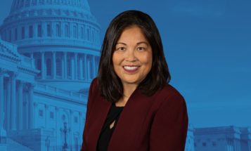 Julie Su in the foreground with U.S. government buildings in the background with a blue overlay