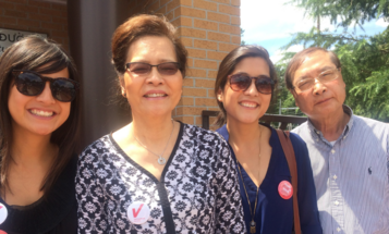A smiling Vietnamese family stands together outdoors, with two women wearing "Vietnamese American Votes" stickers, symbolizing their participation in the voting process. The image reflects pride and unity, emphasizing the role of community support in encouraging voter participation.
