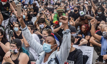 Image accompanying an opinion article titled 'Progressives Won’t Help the Working Class by Abandoning Marginalized Groups' by Taifa Butler and Carol Lautier, published by Demos. It shows a crowd of demonstrators sitting closely together, many wearing masks and raising fists in a sign of solidarity.