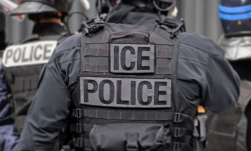 An ICE officer photographed from behind in tactical gear, with "ICE" and "ICE POLICE" patches across the back of a black vest.