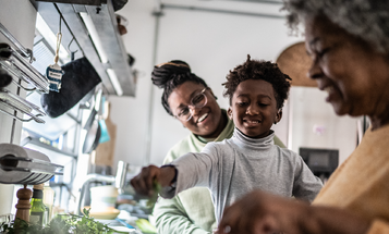 A grandmother, mother, and young boy prepare a meal together in a home kitchen.