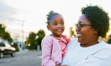 A smiling Black woman in glasses holds a young girl on her hip while standing outdoors on a residential street at sunset.