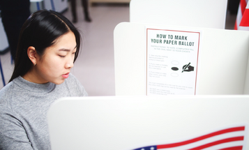 A young woman marks a paper ballot at a voting booth. A posted sign beside her reads "How to mark your paper ballot."