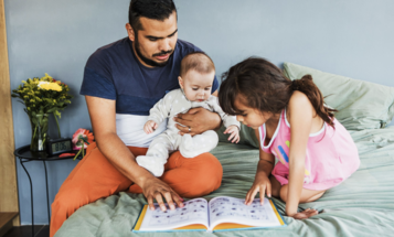 A father reads a picture book on a bed with a baby and a young girl.
