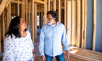 A woman and man smile at each other as they stand inside a house under construction, surrounded by exposed wood framing.