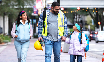 A man in a reflective safety vest walks down a city sidewalk holding hands with a young girl, a hard hat and thermos in his other hand, while an older girl walks alongside them.