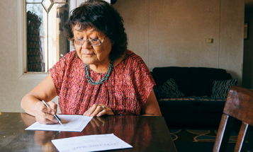 An older woman sits at her dining table filling out a mail-in ballot with a pen, an official election envelope in front of her.