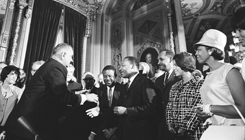 Historic black and white photograph of President Lyndon Johnson signing civil rights legislation surrounded by MLK Jr. and other civil rights leaders and officials in an ornate government building
