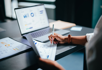 Person writing on a financial report in front of a laptop