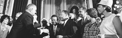 Historic black and white photograph of President Lyndon Johnson signing civil rights legislation surrounded by MLK Jr. and other civil rights leaders and officials in an ornate government building