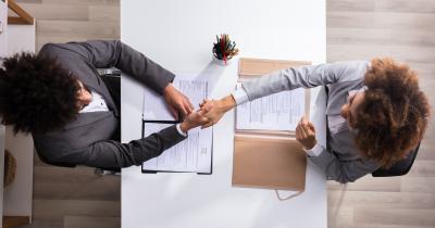 Two people shaking hands across a table in an interview