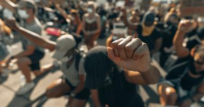 Black, brown, and white protestors knee with their heads down and their fists in the air