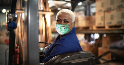 Black warehouse worker wearing mask operating forklift