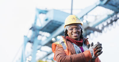 Smiling Black construction worker with a yellow hard hat and safety vest in front of a construction crane