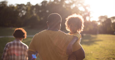 Backlit image of a Black family walking towards the setting sun across an open lawn with a toddler looking over their father's shoulder toward the camera