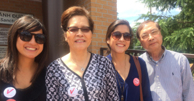 A smiling Vietnamese family stands together outdoors, with two women wearing "Vietnamese American Votes" stickers, symbolizing their participation in the voting process. The image reflects pride and unity, emphasizing the role of community support in encouraging voter participation.