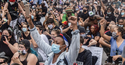 Image accompanying an opinion article titled 'Progressives Won’t Help the Working Class by Abandoning Marginalized Groups' by Taifa Butler and Carol Lautier, published by Demos. It shows a crowd of demonstrators sitting closely together, many wearing masks and raising fists in a sign of solidarity.