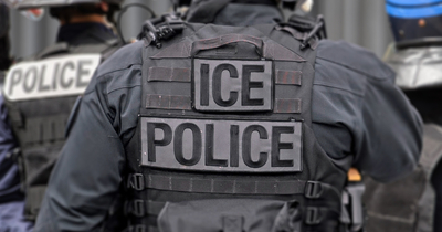 An ICE officer photographed from behind in tactical gear, with "ICE" and "ICE POLICE" patches across the back of a black vest.
