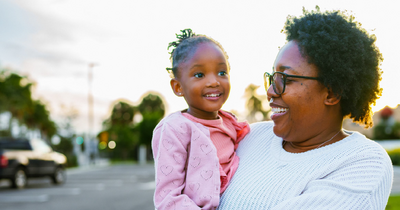 A smiling Black woman in glasses holds a young girl on her hip while standing outdoors on a residential street at sunset.