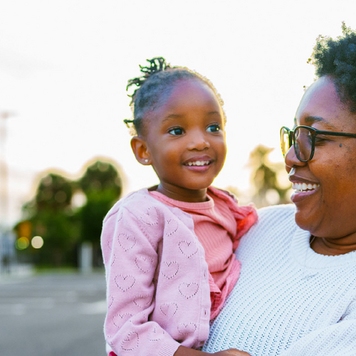 A smiling Black woman in glasses holds a young girl on her hip while standing outdoors on a residential street at sunset.