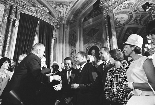 Historic black and white photograph of President Lyndon Johnson signing civil rights legislation surrounded by MLK Jr. and other civil rights leaders and officials in an ornate government building